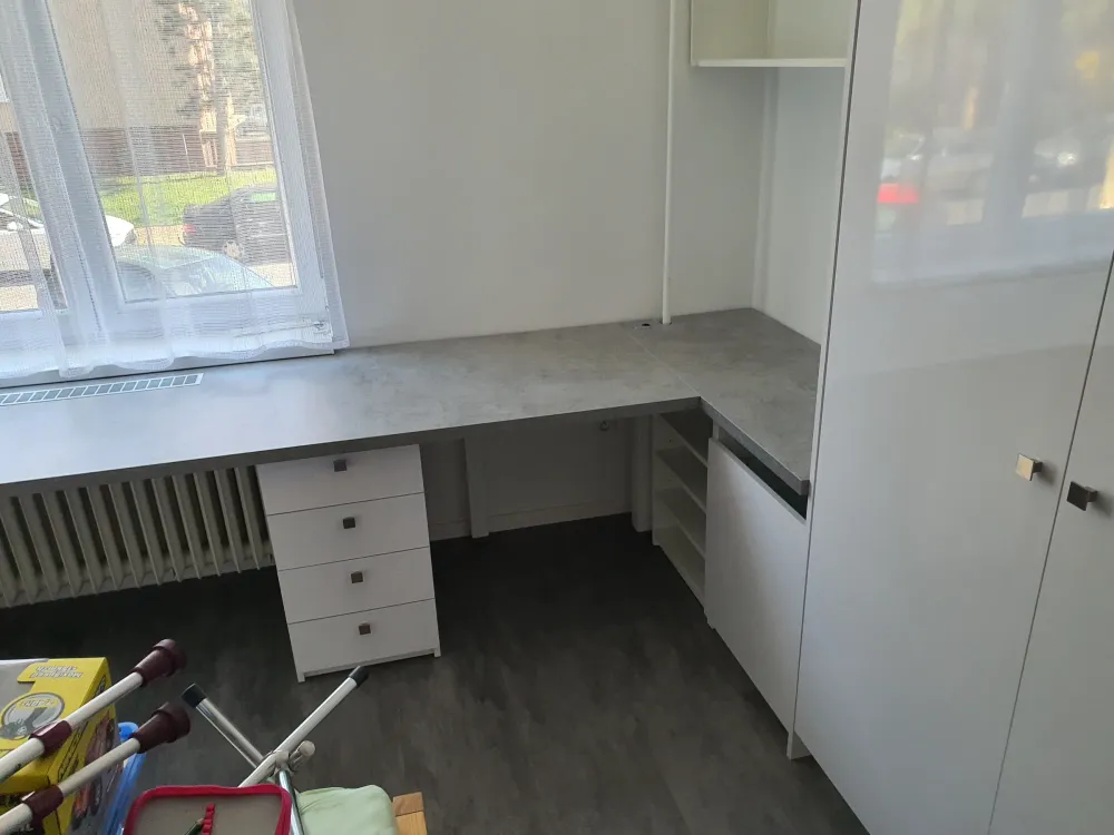 Rohový bílý psací stůl pod oknem s&nbsp;kontejnerem a&nbsp;bílou skříní