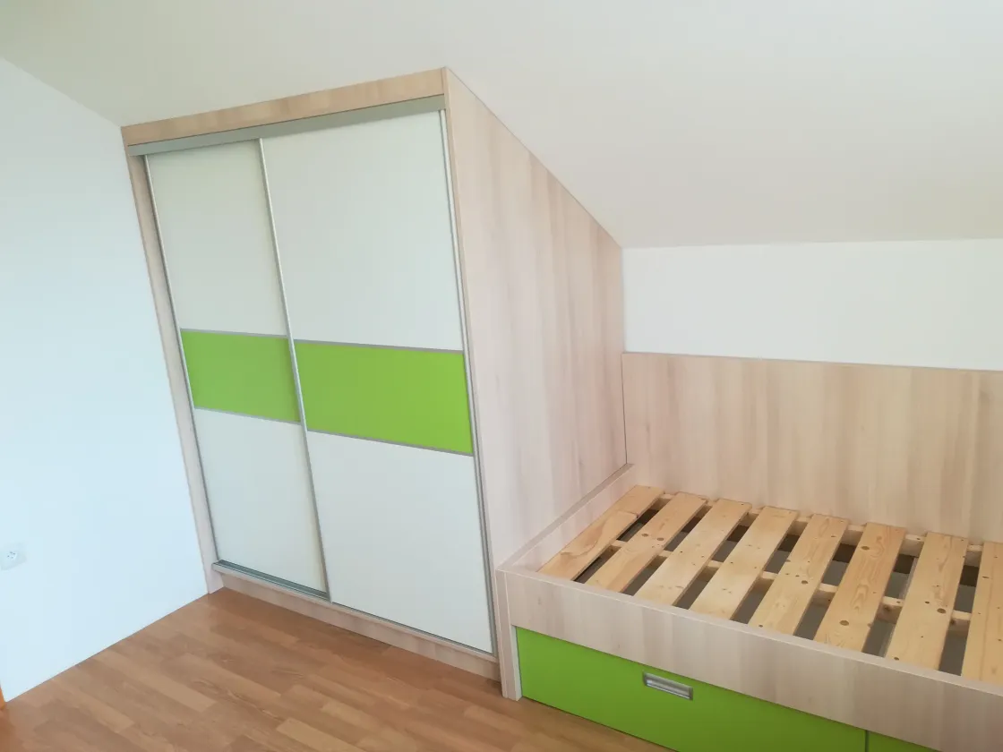 Rošt postele pod šikminou, zelené šuplíky a&nbsp;psací stůl u&nbsp;okna