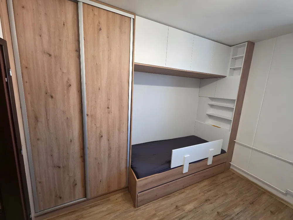 Posuvné dveře šatní skříně v&nbsp;dekoru dubu, vedle postel se zábranou a&nbsp;bílé nástavce nad ní