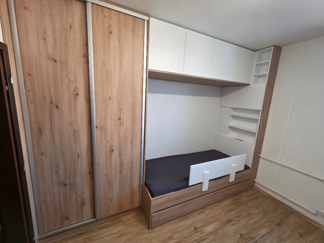 Posuvné dveře šatní skříně v&nbsp;dekoru dubu, vedle postel se zábranou a&nbsp;bílé nástavce nad ní