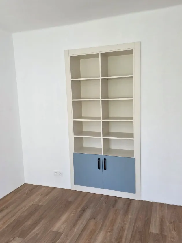 Bílá knihovna vestavěná do niky ve zdi, dole modrá dvířka s&nbsp;černými úchytkami