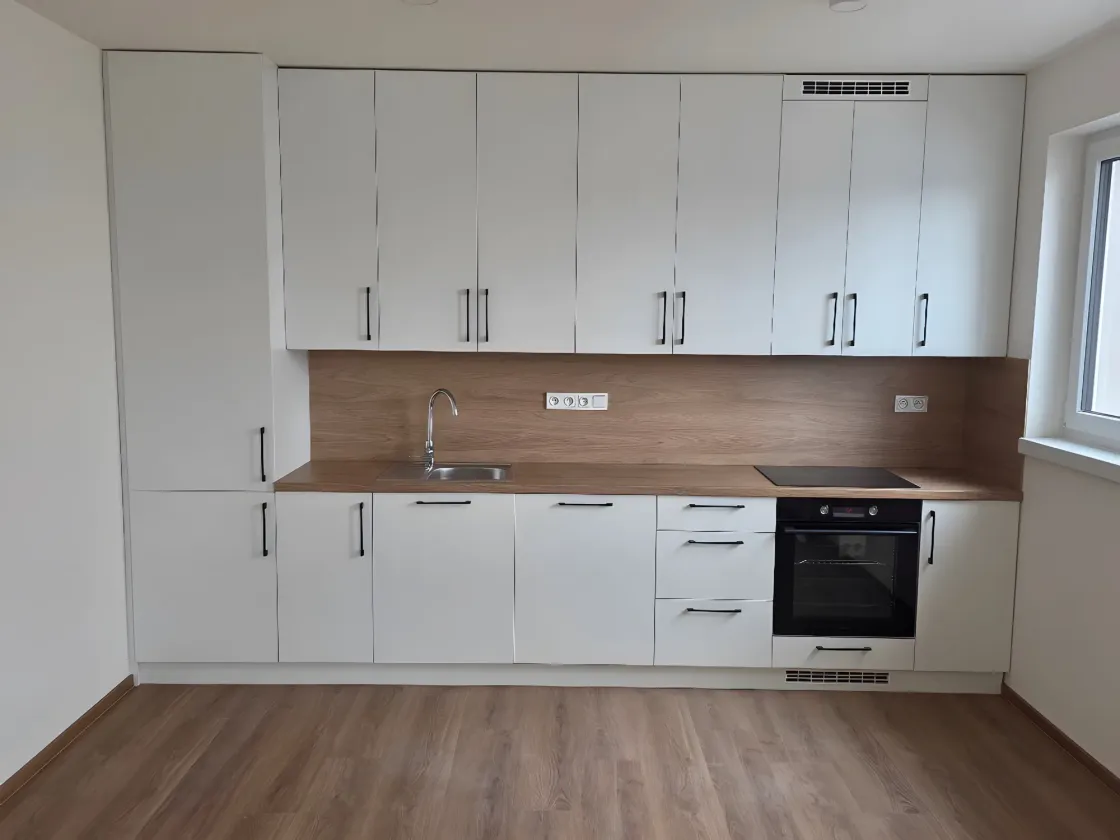 Rovná bílá linka s&nbsp;černými úchytkami, deska a&nbsp;obklad v&nbsp;dekoru dubu, trouba pod varnou deskou