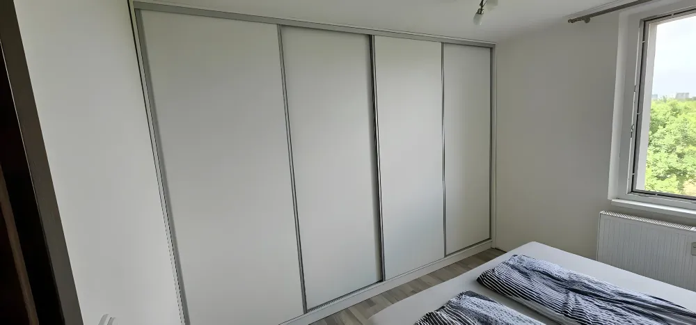 Bílá šatní skříň s&nbsp;posuvnými dveřmi za postelí