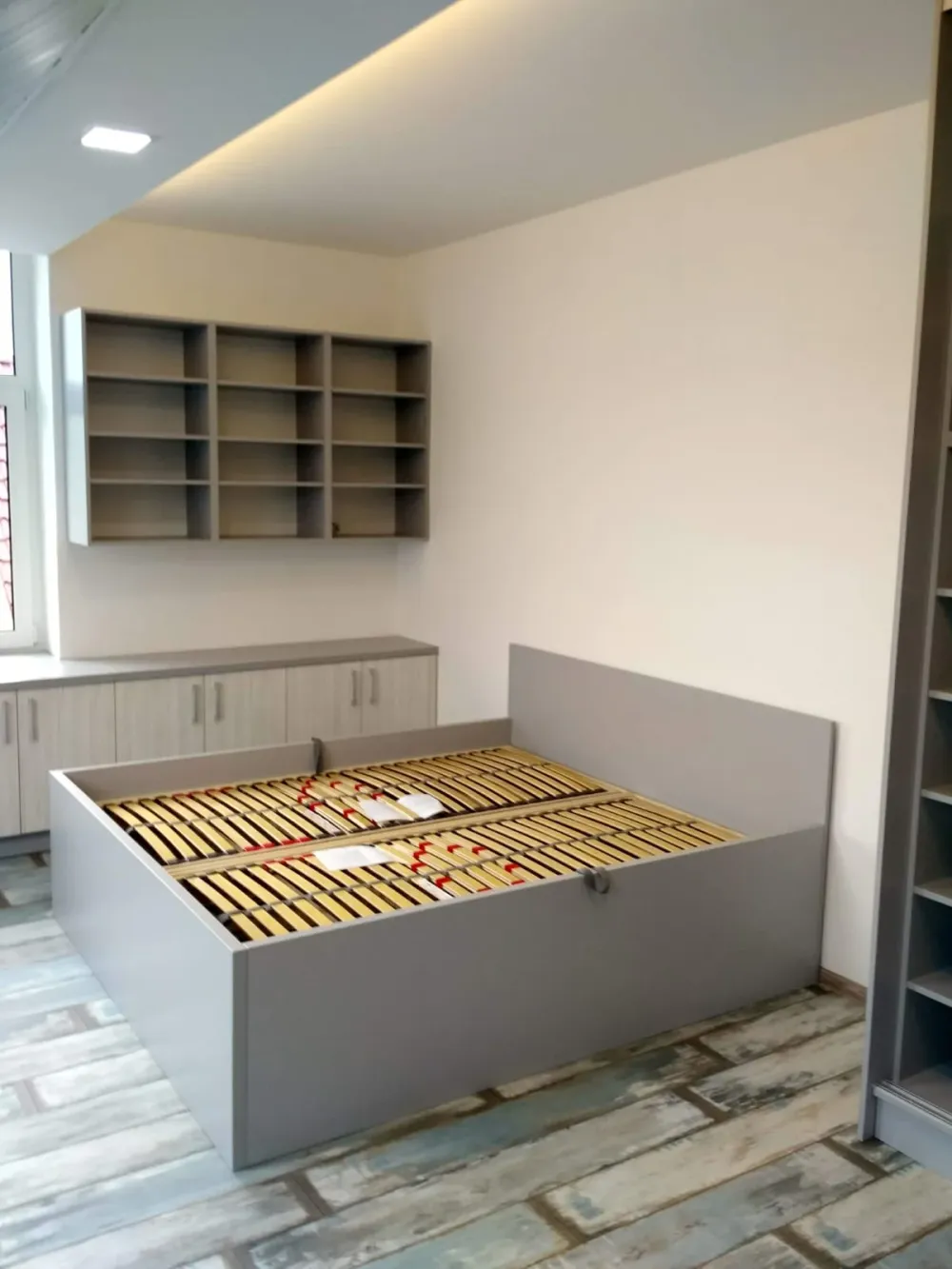 Šedá manželská postel s&nbsp;roštem v&nbsp;podkroví, za ní police na stěně a&nbsp;skříňky pod oknem