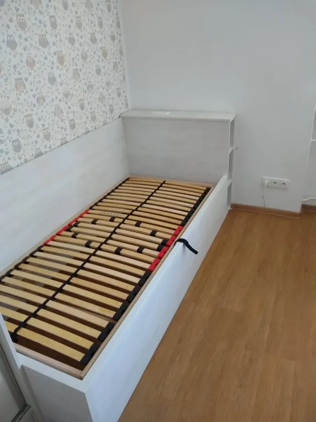 Bílá jednolůžková postel s&nbsp;roštem a&nbsp;policemi v&nbsp;čele u&nbsp;stěny s&nbsp;tapetou se sovami
