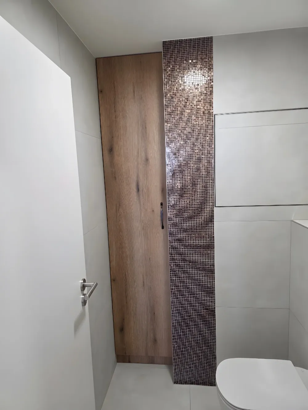 Úzká vysoká skříň v&nbsp;dekoru dubu vestavěná vedle obkladu se sloupcem mozaiky na WC