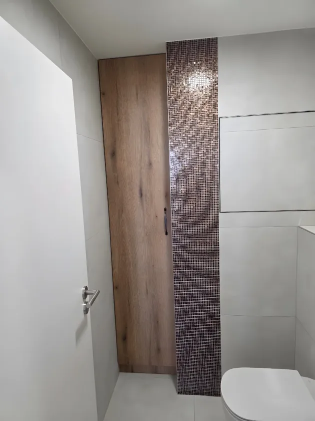 Úzká vysoká skříň v&nbsp;dekoru dubu vestavěná vedle obkladu se sloupcem mozaiky na WC